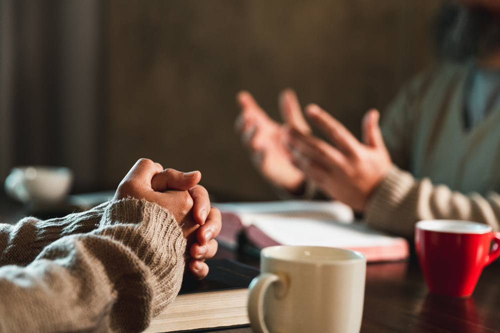 People raising hands and folding in prayer at table with Bibles and coffee cups laid out
